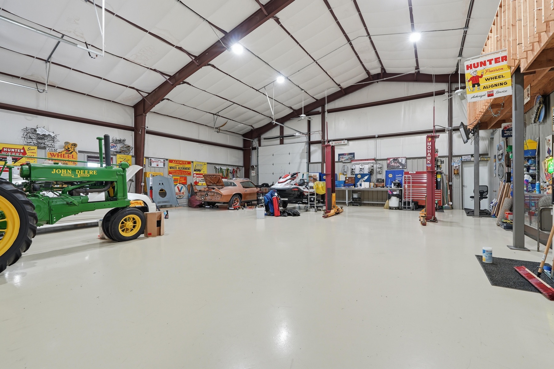 Interior of the 7,500 sq ft red-iron commercial shop with polished concrete floor, a vintage green John Deere tractor on the left, a project muscle car under work on the right, LED lighting suspended from steel trusses overhead, and a Hunter wheel alignment sign on the back wall