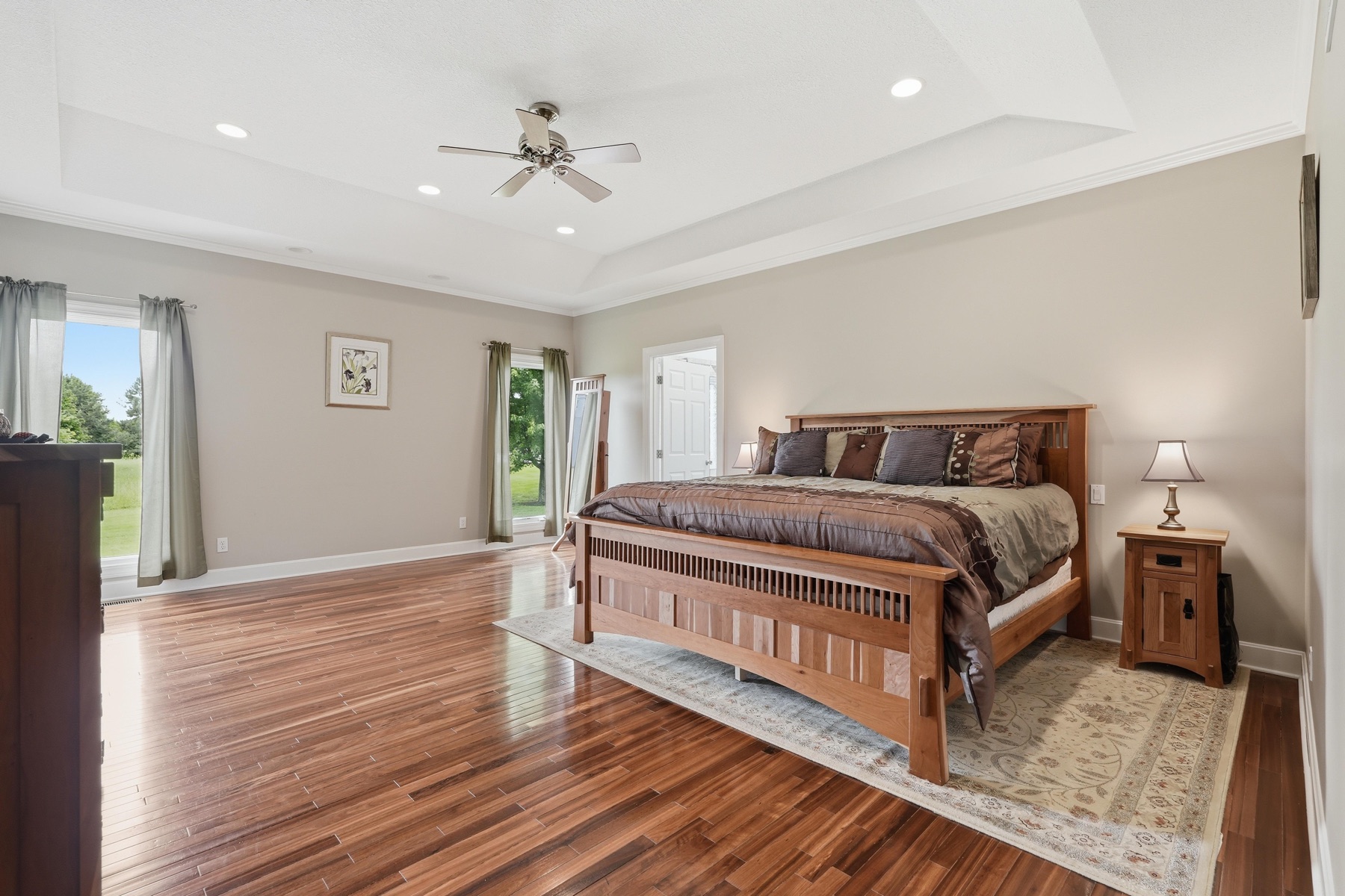 Primary bedroom with coffered tray ceiling, ceiling fan, mission-style king bed with carved headboard, exotic hardwood floors stretching wall to wall, and windows letting green light across the room