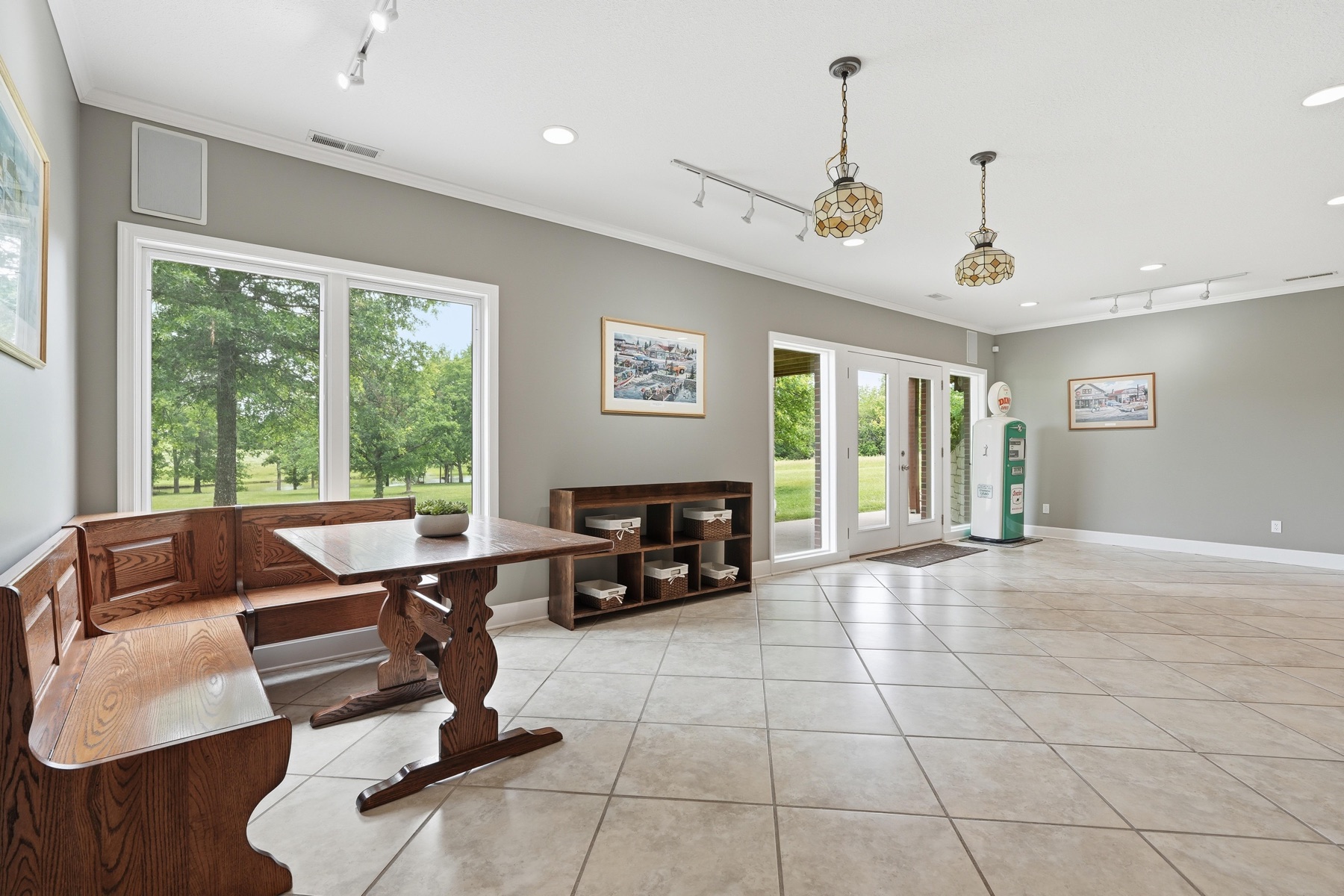 Sun room entry vestibule with French doors looking onto green Kansas pasture, a restored vintage green Sinclair Dino gas pump standing as a collector piece beside the doors, a built-in window bench seat to one side, and a track-lit ceiling overhead