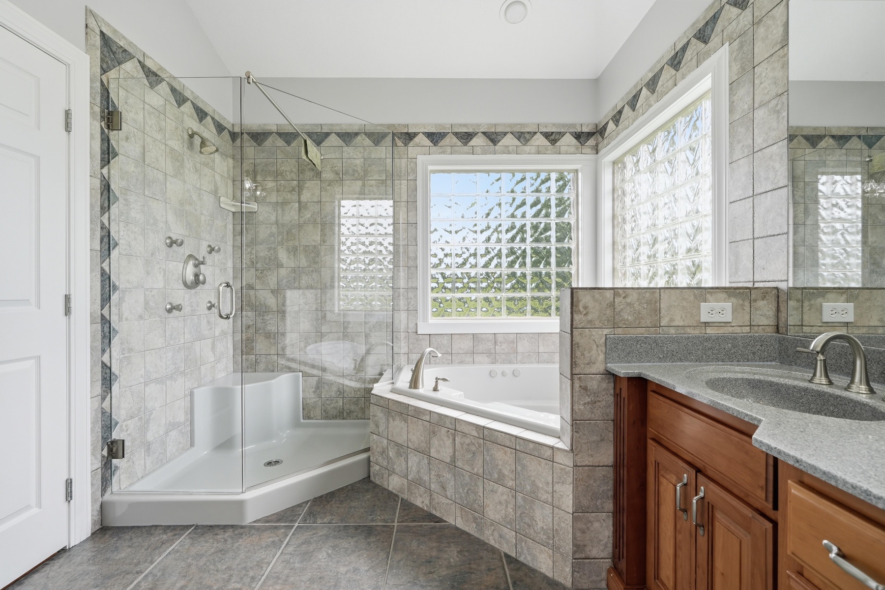 Primary bathroom showing the corner jacuzzi tub framed in tile, a glass block window flooding the corner with diffused light, a walk-in shower stall with chevron tile detail and frameless glass door, and a cherry-wood vanity to the right