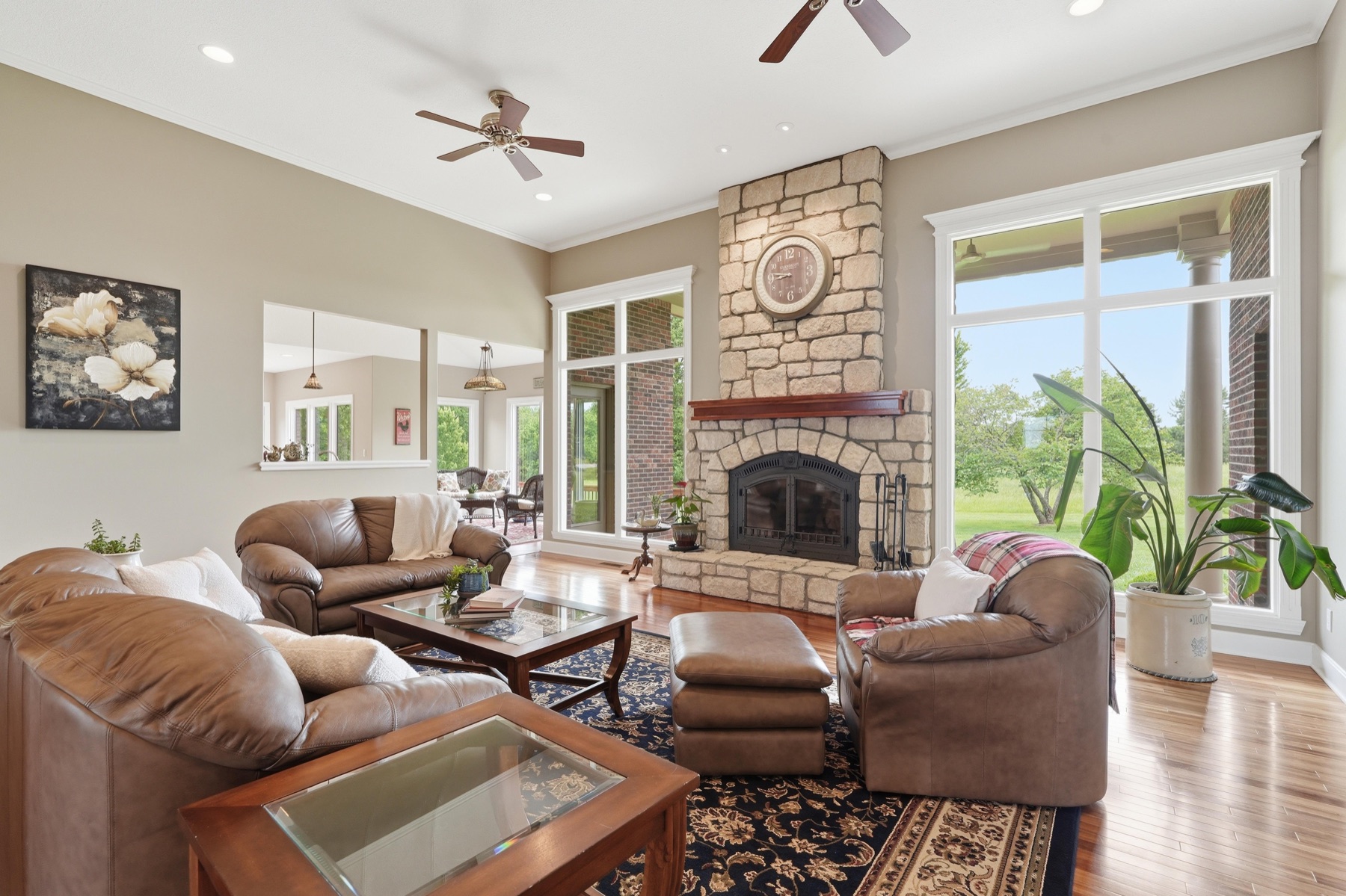 Great room at 26619 K-68 Hwy with stacked stone fireplace centered between transom windows, two large brown leather sofas flanking a glass-top coffee table, tropical plants by the window, and two ceiling fans hanging from the vaulted ceiling
