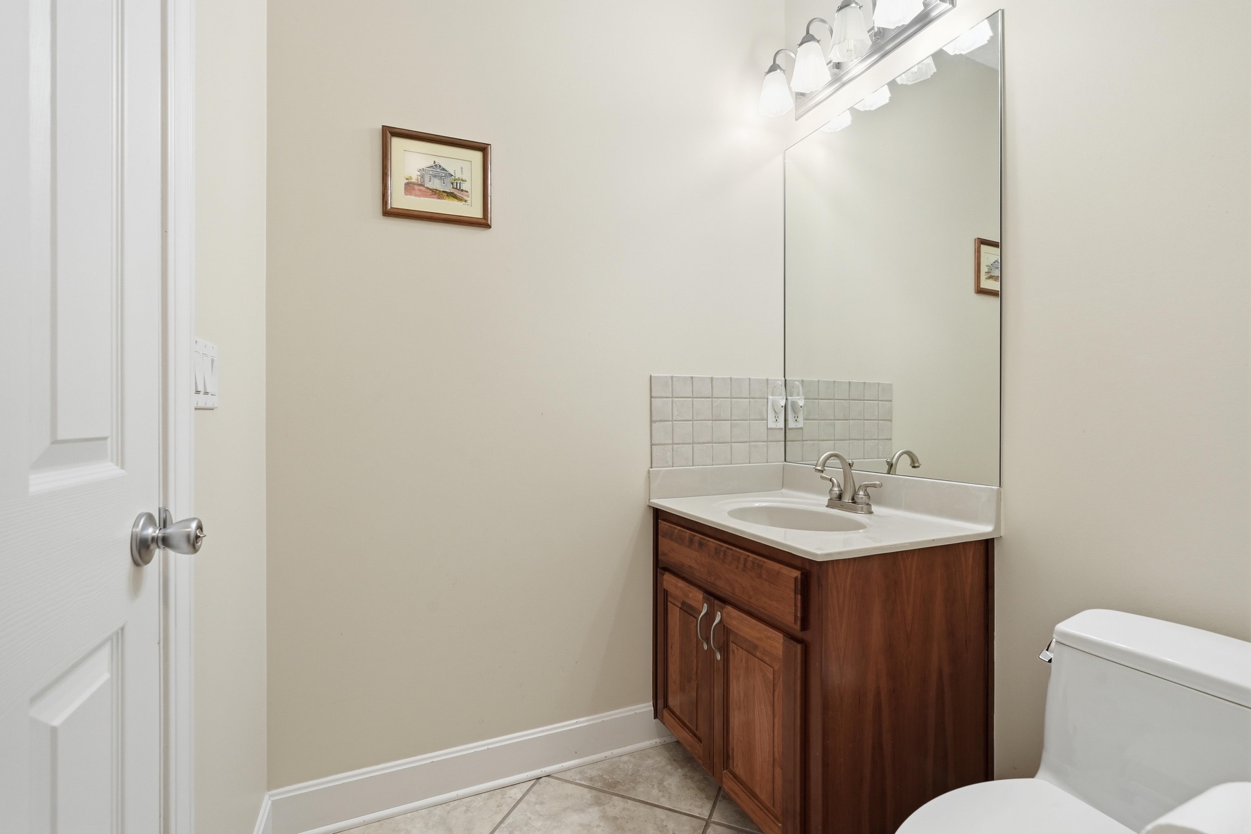Powder room with a cherry-wood vanity, single sink with brushed nickel faucet, four-bulb vanity light over a wide mirror, a small framed watercolor of a country house on the wall, and tile floor
