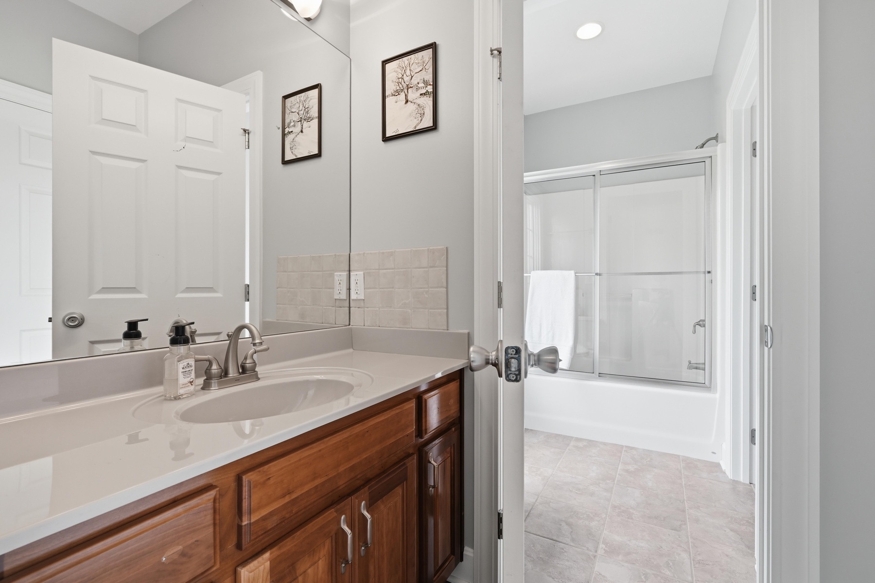 Hall bathroom with two framed prints over a long wood vanity with single sink, a glass-enclosed tub-shower combo with sliding doors, mirror reflecting the door, and tile flooring