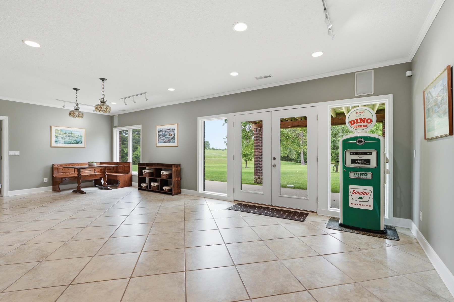 Open tile-floor entry hall with track lighting, a pair of pendant chandeliers above an antique sideboard, French doors opening onto the back deck, and the green vintage Sinclair Dino gas pump standing prominently between the doors