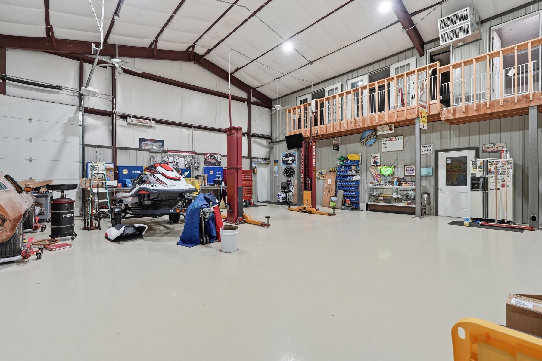 Wider angle of the shop interior showing the mezzanine office built across the back wall with a wood railing, a personal watercraft on a lift to the left, a window AC unit visible upstairs, a side overhead door, and steel truss roof construction overhead