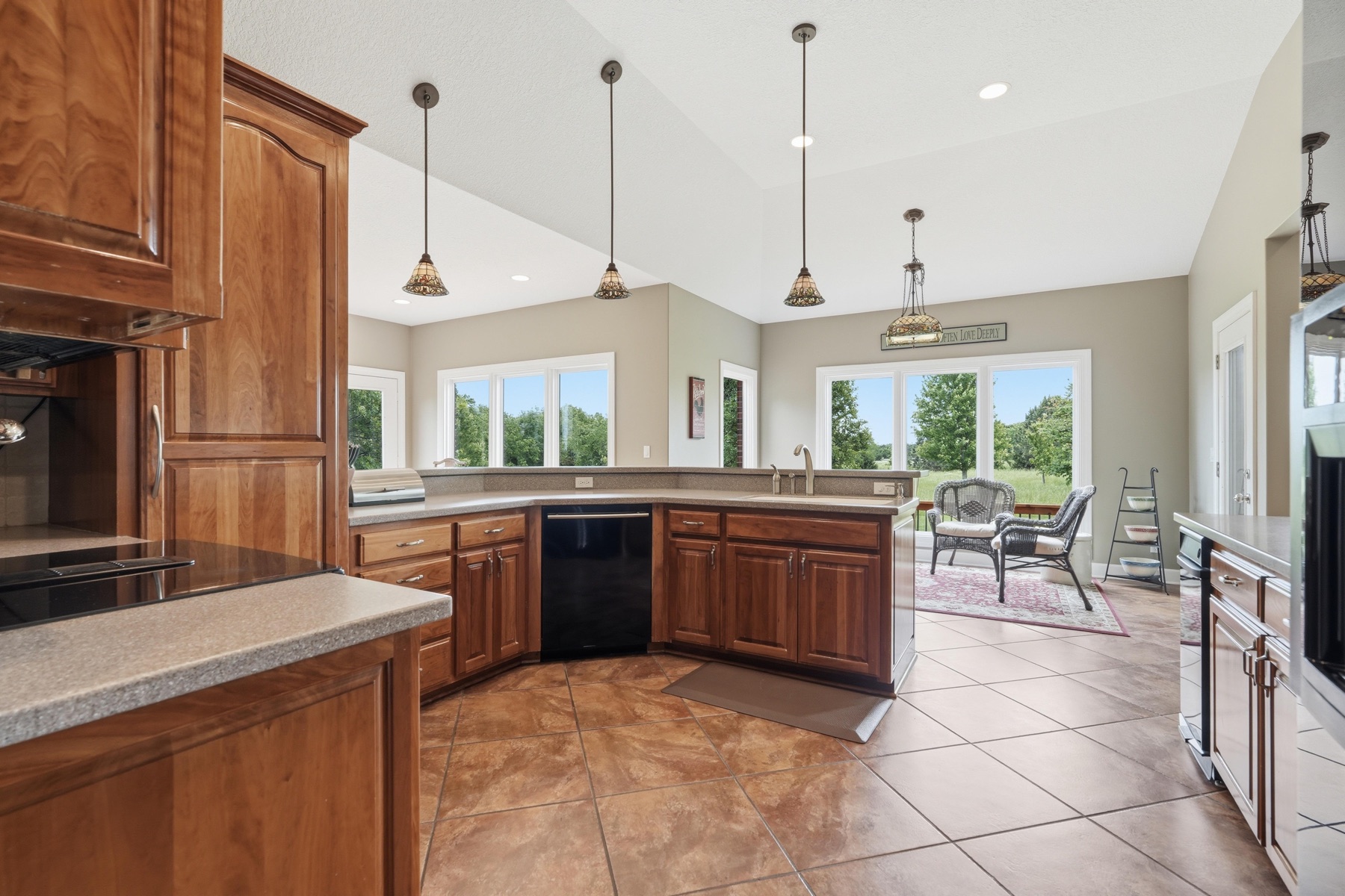 Kitchen with custom cherry-wood cabinetry, a long peninsula with three pendant lights overhead in copper and stained glass, a black dishwasher, vaulted ceiling, and a clear sightline through to the breakfast nook and back windows
