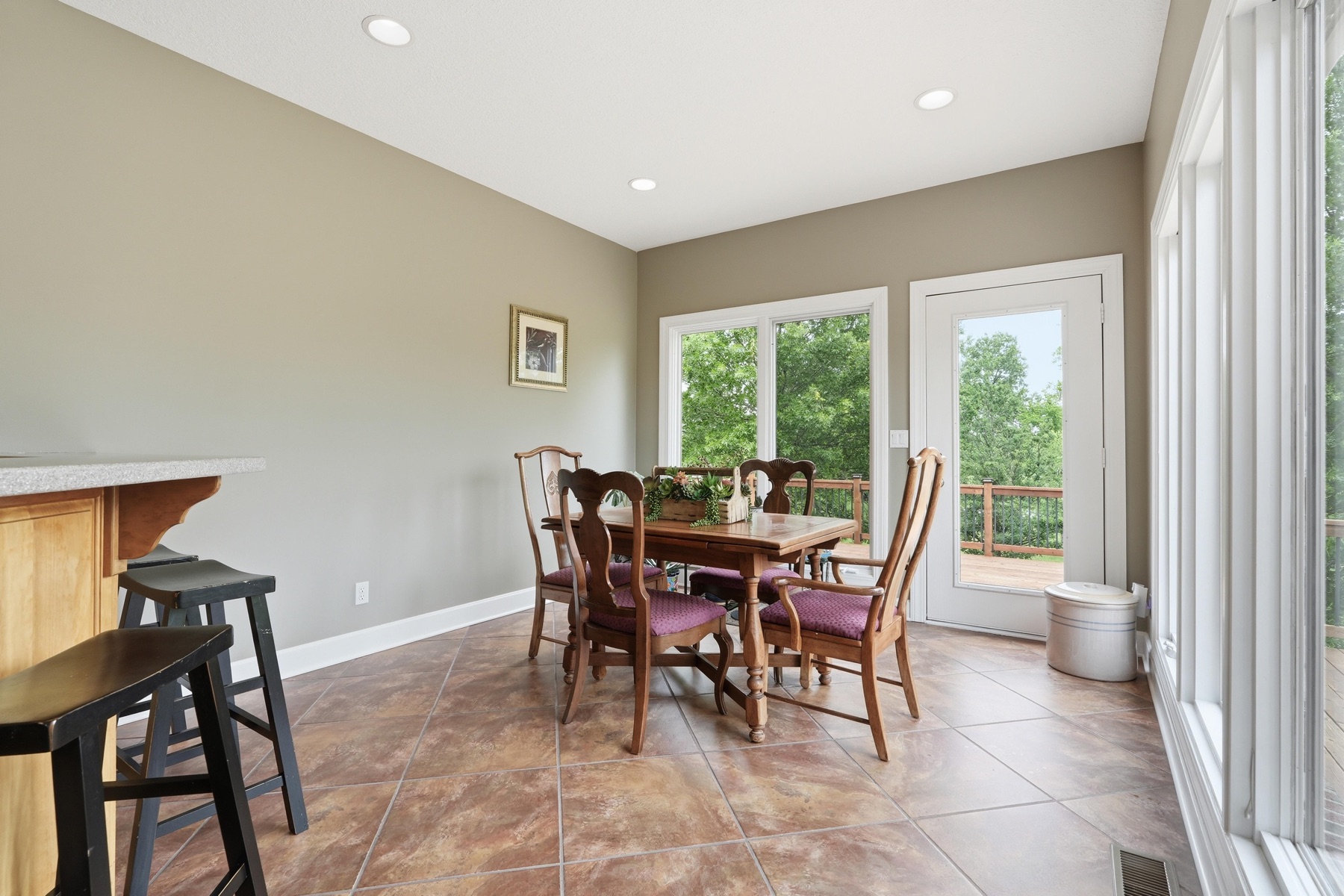 Breakfast dining nook with a small wood table and four high-backed chairs, French door leading out to the cedar deck and the green tree line beyond, large picture windows on two walls, and warm tile floors throughout