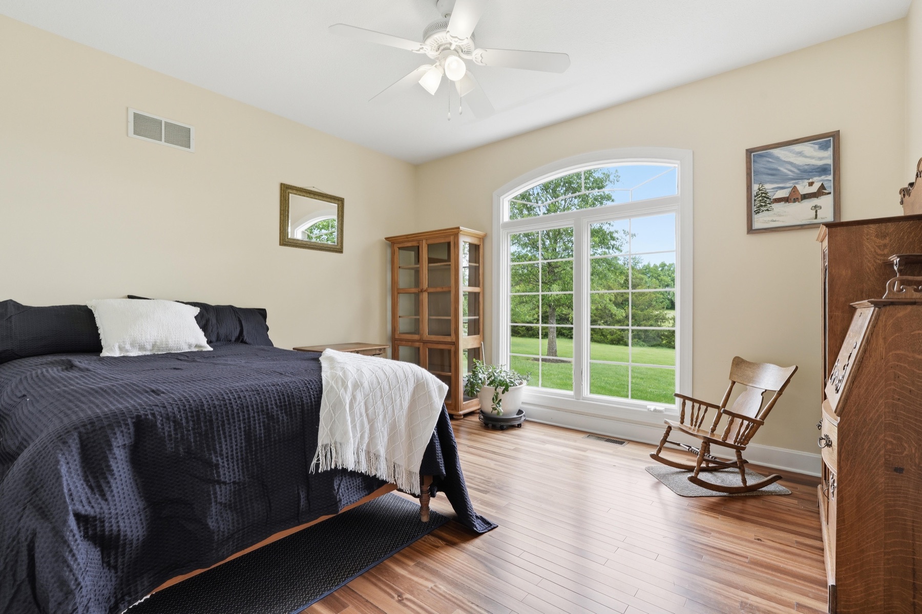 Secondary bedroom with vaulted ceiling, an arched picture window overlooking the green front lawn, a tall wood armoire with diamond-paned glass doors, a wooden rocking chair, and warm hardwood floors