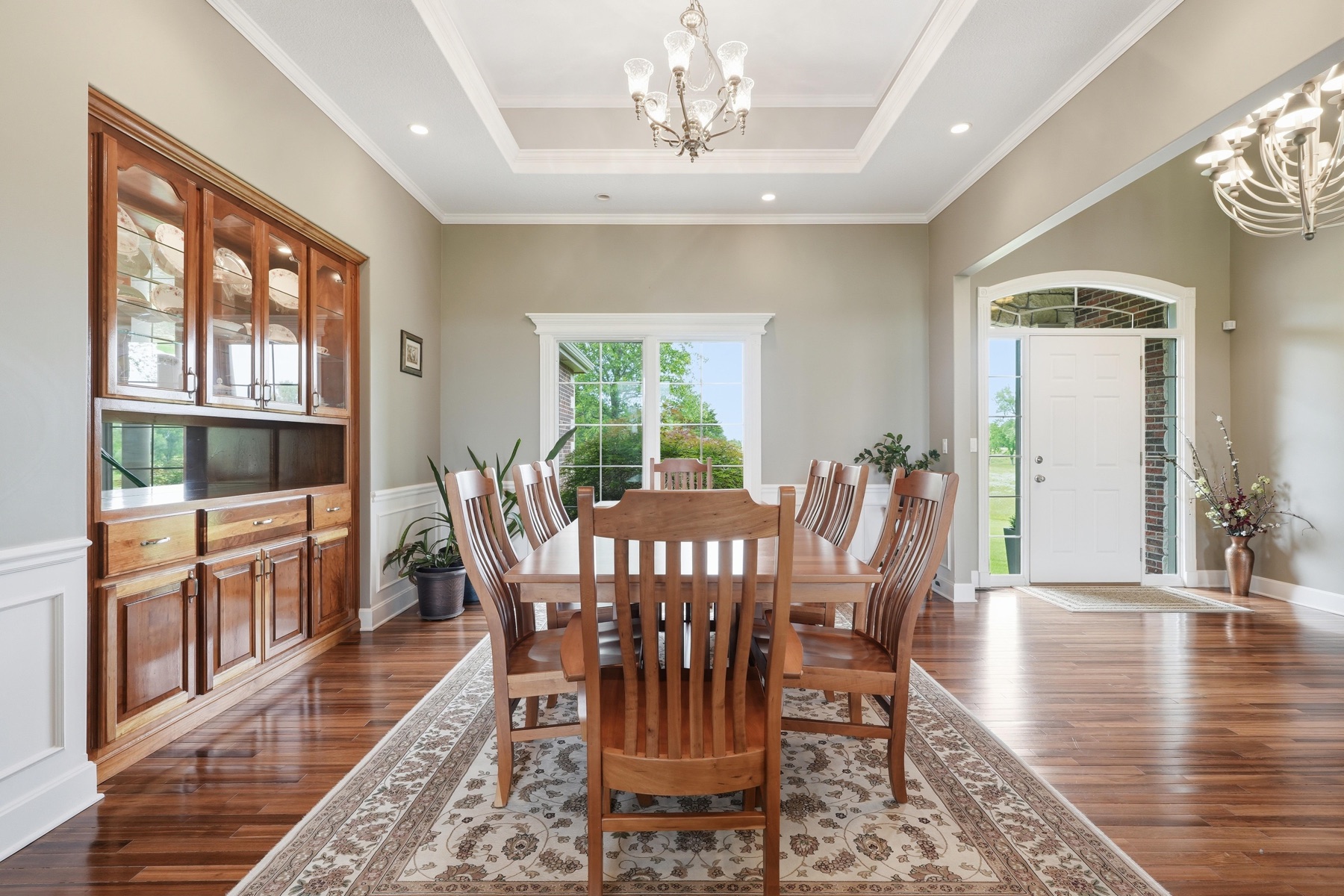 Formal dining room with double-tray ceiling, two crystal chandeliers, an eight-chair mission-style wood table on a cream Persian rug, a tall cherry-wood china cabinet built into the wall, and windows letting in green light from the side yard