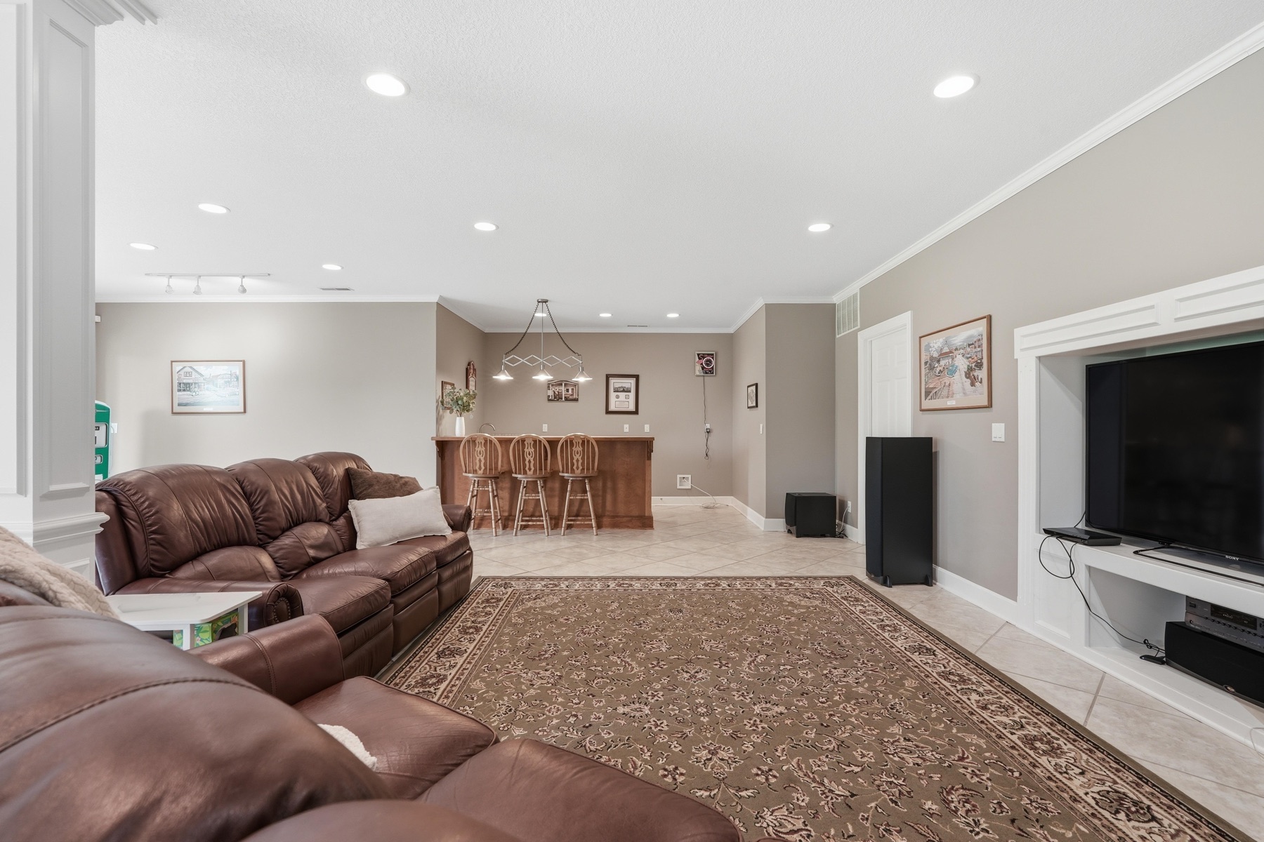 Lower-level family room with two brown leather sectional sofas on a tan Persian rug, a wall-mounted flat-screen TV, a wet-bar pass-through to the left, tile flooring, and a built-in entertainment cabinet to the right