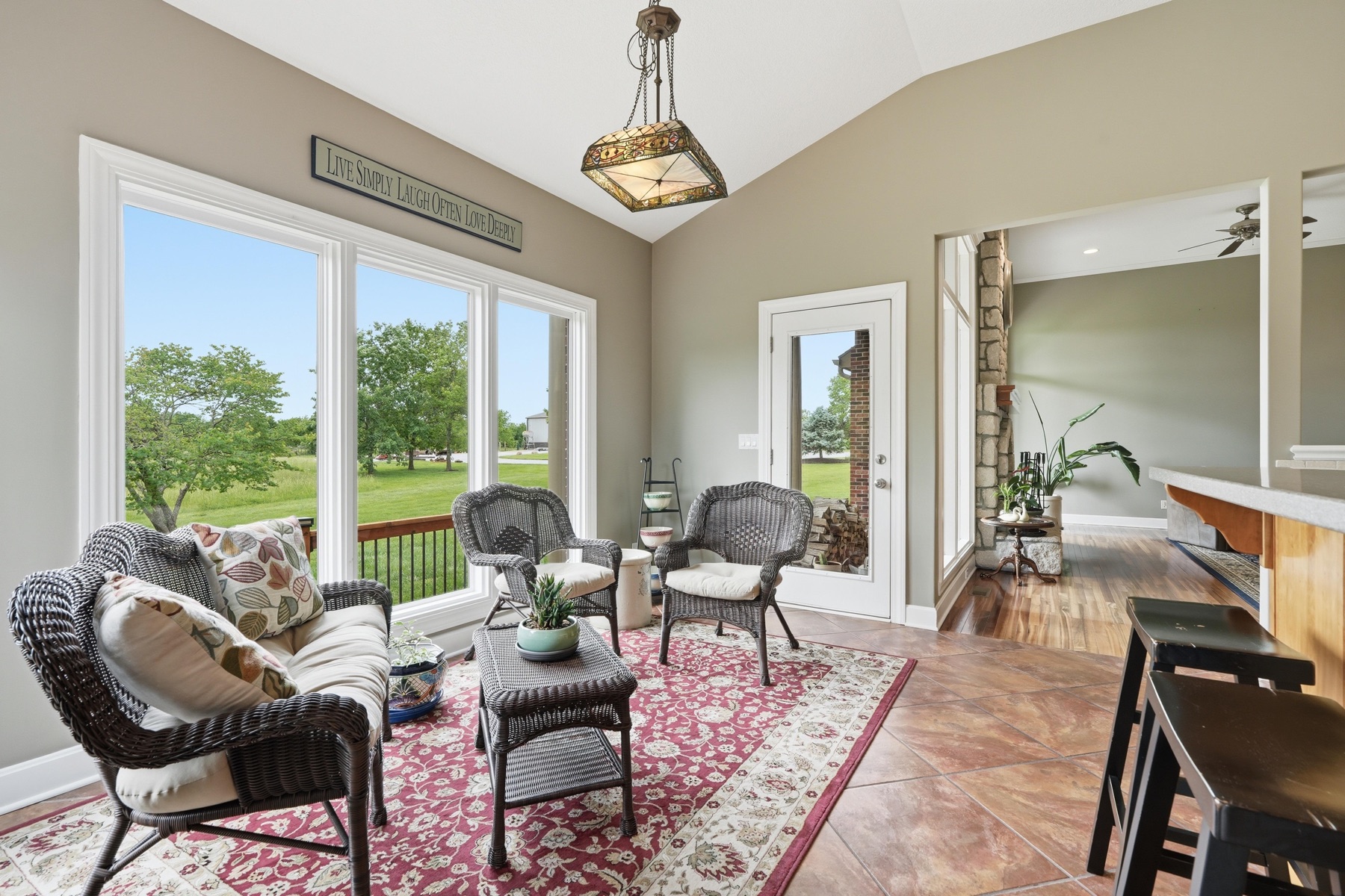 Sun room with three walls of windows letting in green light, wicker seating with patterned cushions on a Persian rug, a stained-glass pendant lantern overhead, and a sign on the wall reading Live Simply Laugh Often Love Deeply