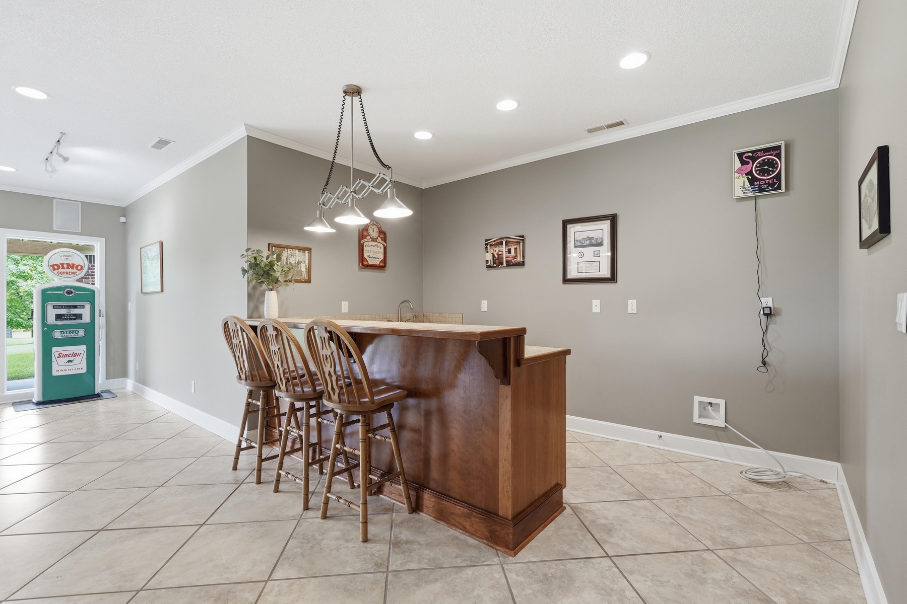 Wet-bar area with three high stools at a curved cherry-wood bar, a Sinclair-style pendant fixture overhead, framed posters and vintage gas station signage on the walls, a clock styled like a fuel gauge, and the green Sinclair pump just visible to the left