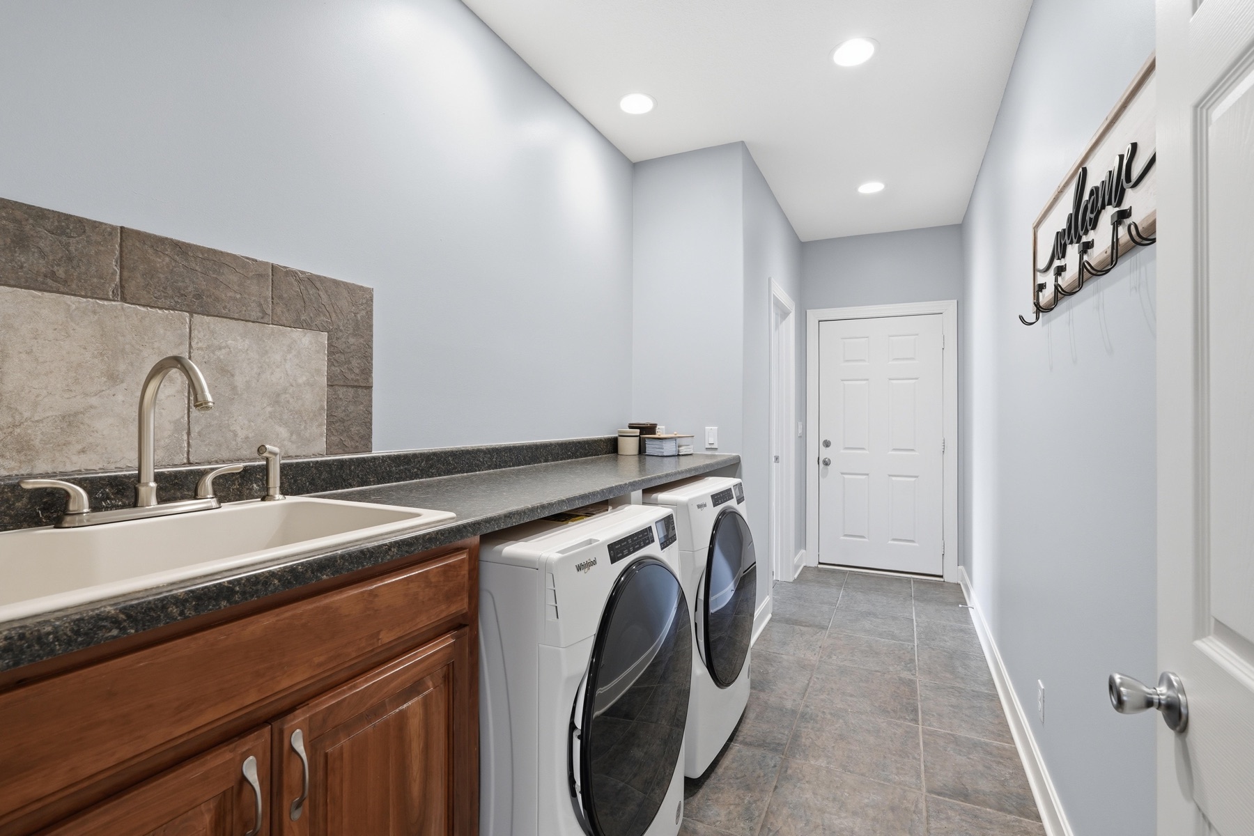 Main-level laundry room with a deep utility sink under a tiled backsplash, side-by-side high-efficiency front-loading washer and dryer in white, custom cabinetry above for storage, and a door at the back leading to the rest of the home