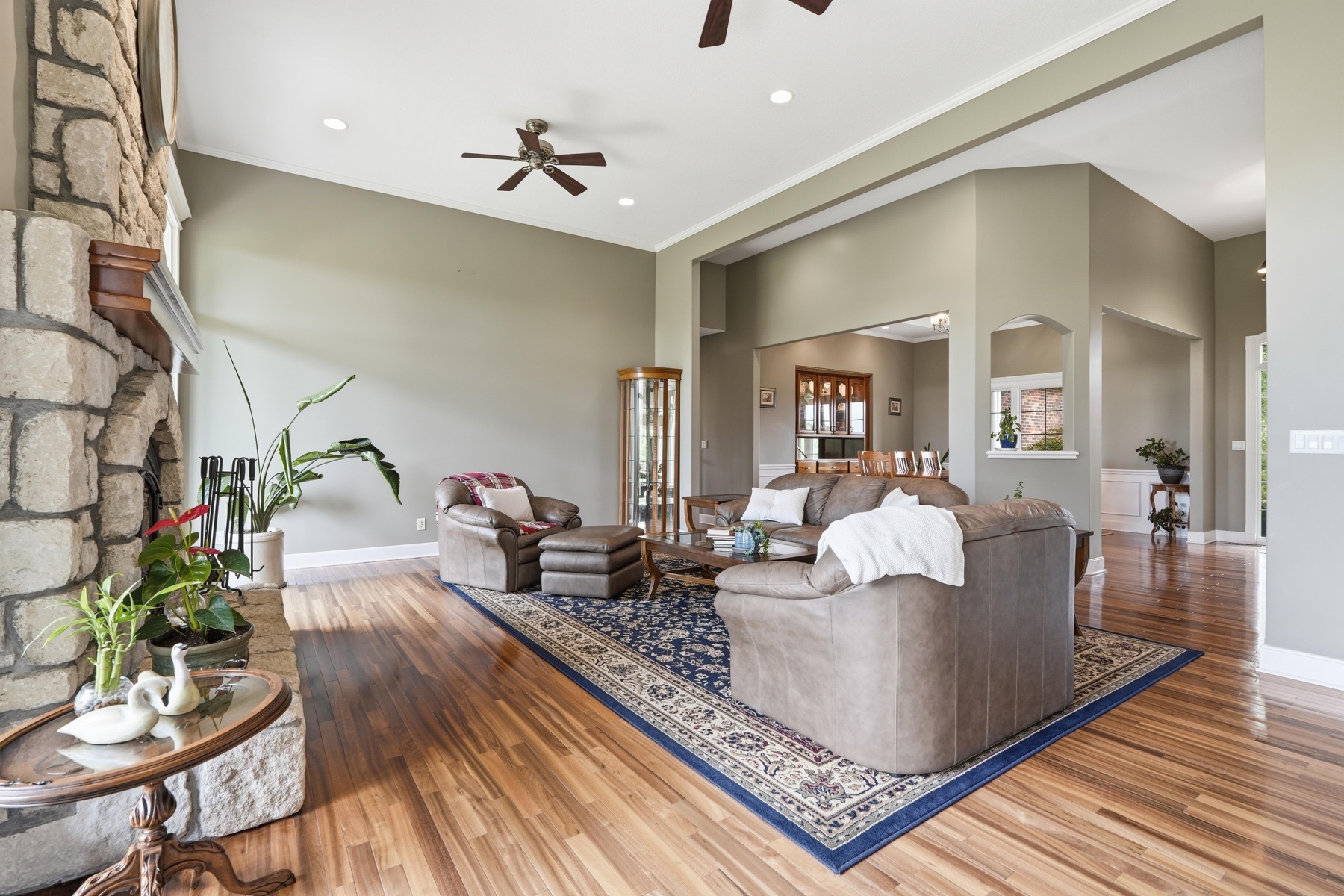 Great room from a third angle showing the stacked-stone fireplace on the left, two leather chairs and an ottoman on a deep blue Persian rug, openings into the dining room and kitchen on the right, and exotic hardwood floors stretching throughout