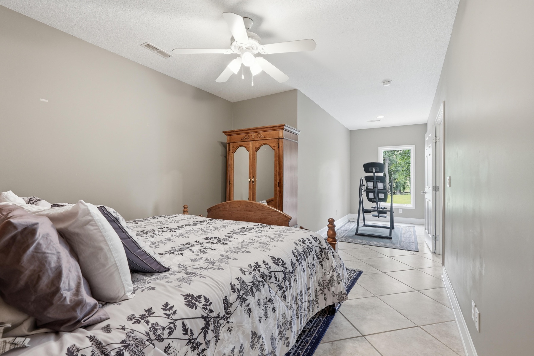 Second bedroom with vaulted ceiling, tile flooring, an antique armoire with diamond-paned glass doors against the back wall, an exercise stair machine in the corner, and a window letting in green light from the yard