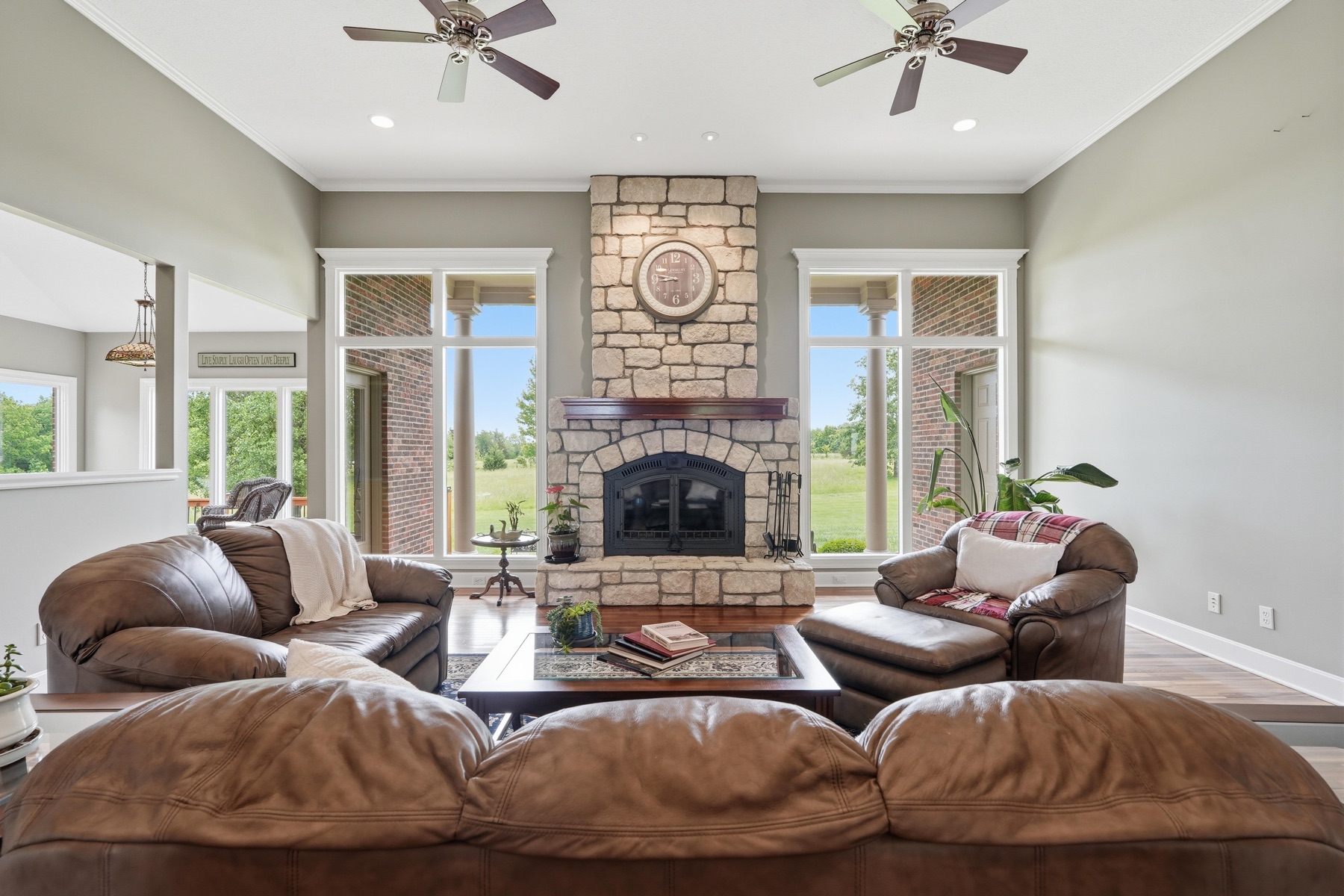 Great room from the main couch perspective looking at the stone fireplace centered between two transom windows, a brown leather sectional in the foreground, two leather armchairs flanking the hearth, two ceiling fans overhead, and hardwood floors throughout