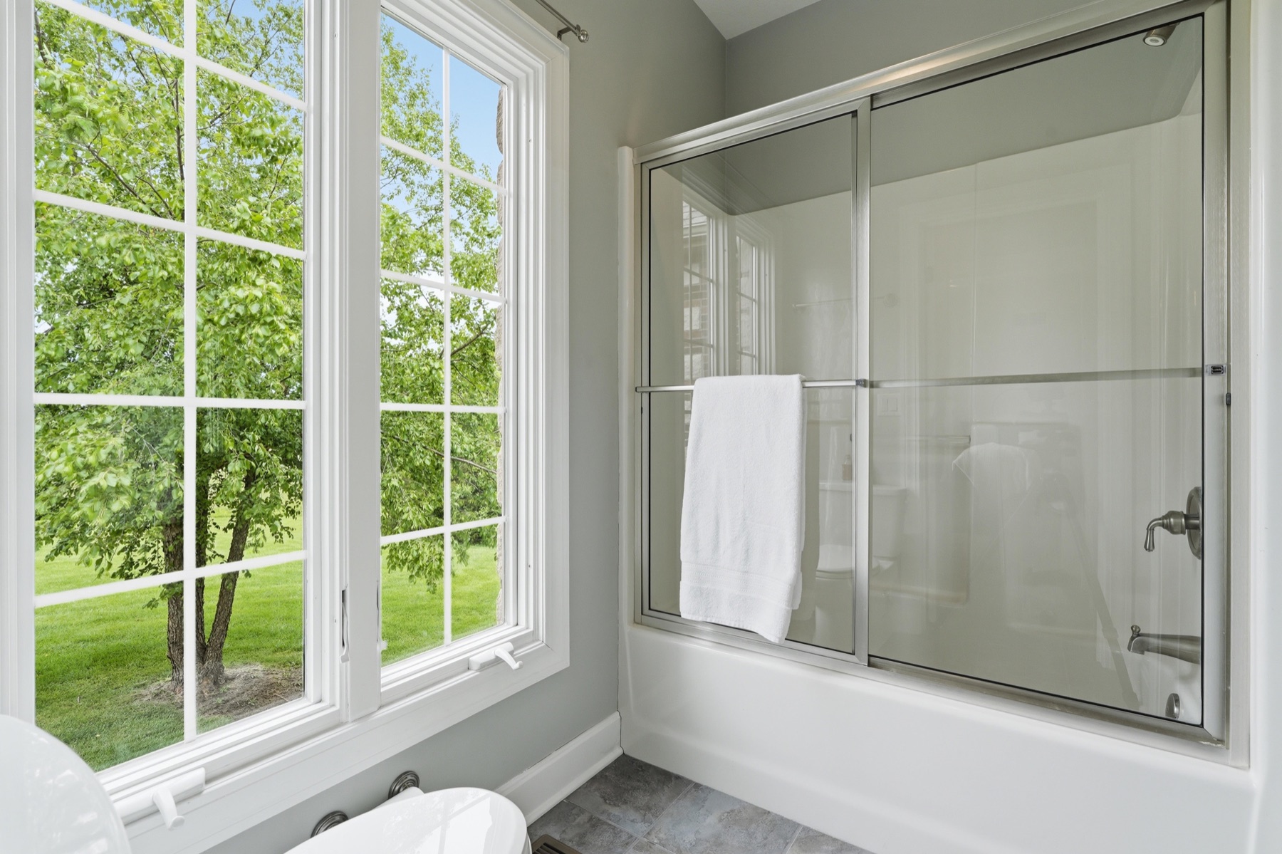 Secondary bathroom showing a tub-shower combo with frameless glass sliding doors, a large picture window with white grid framing the green tree line outside, soft daylight flooding the room, and tile floor underfoot