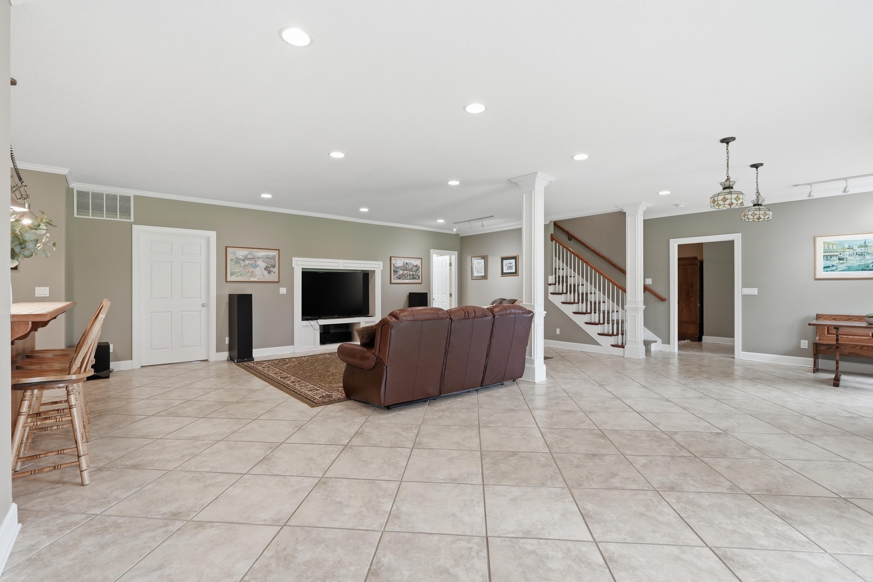 Lower-level family room with brown leather sectional, wall-mounted TV, tile floor, a carpeted staircase leading up to the main level on the right, two columns marking the opening to the rest of the lower level, and recessed lighting throughout