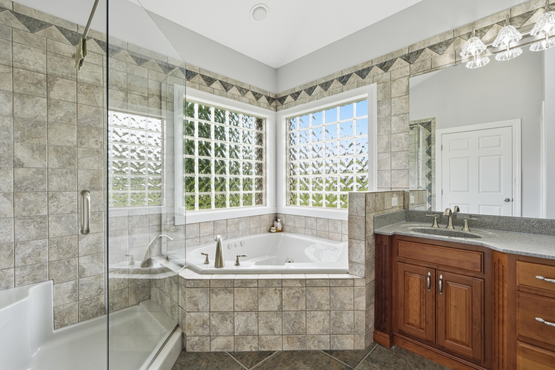 Primary bathroom wide view showing the corner jacuzzi tub framed by tile and a tall glass-block window letting diffused daylight in, the walk-in shower with frameless glass door on the left, the long cherry-wood double-sink vanity on the right, and crown moulding overhead