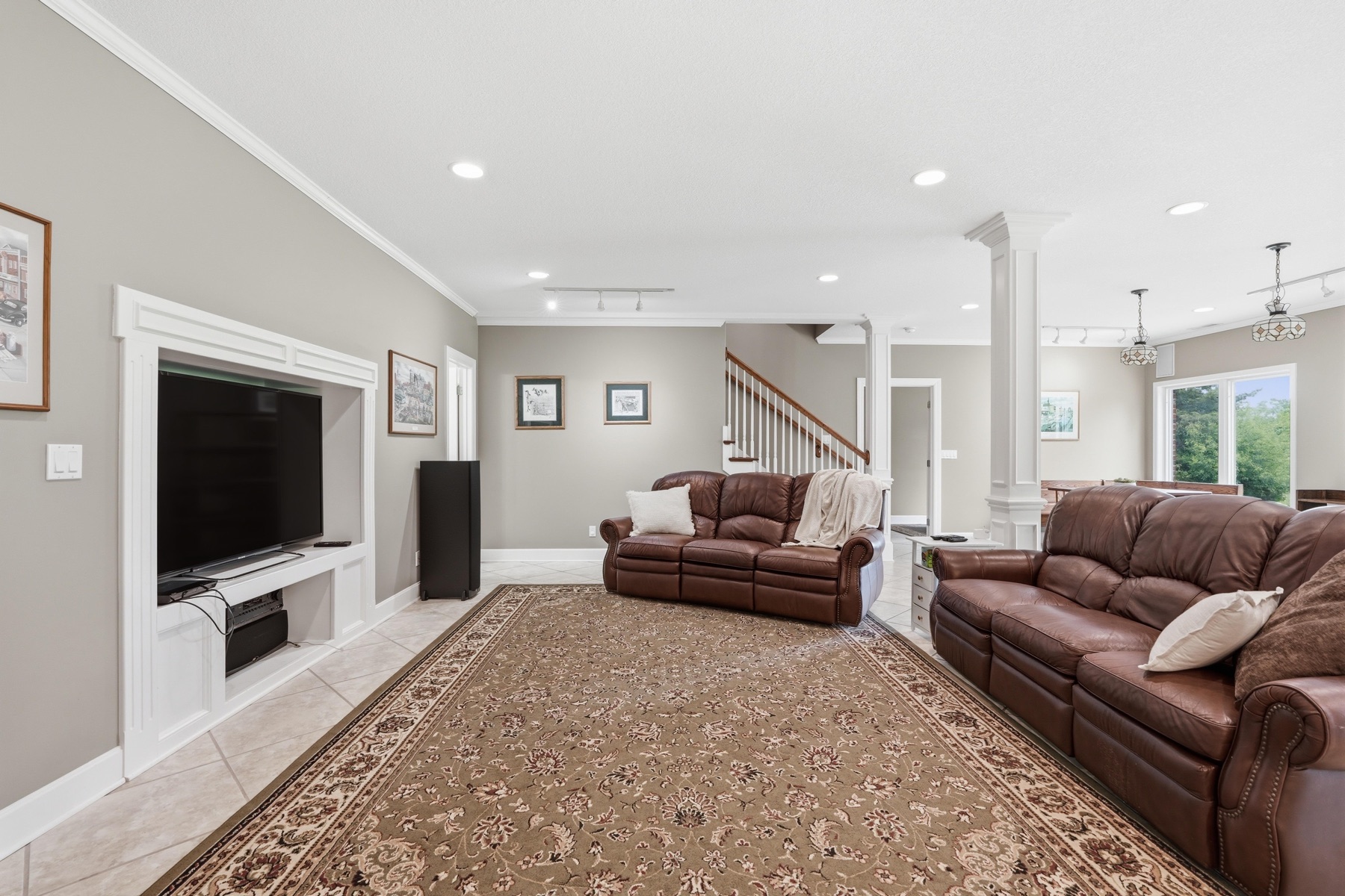 Lower-level family room from a different angle showing two brown leather sofas, a wall-mounted flat-screen TV in a built-in white surround, a vintage speaker tower in the corner, tile floor with a brown Persian rug, and the stairs visible in the background