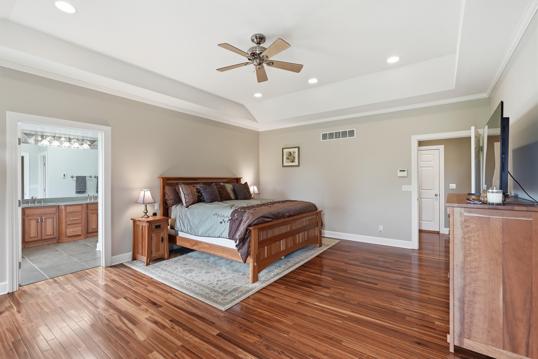 Primary bedroom from the bath door showing the mission-style bed on a wool rug, the tray ceiling with ceiling fan, a doorway to the primary bath visible to the left, a doorway to a walk-in closet on the right, exotic hardwood floors throughout