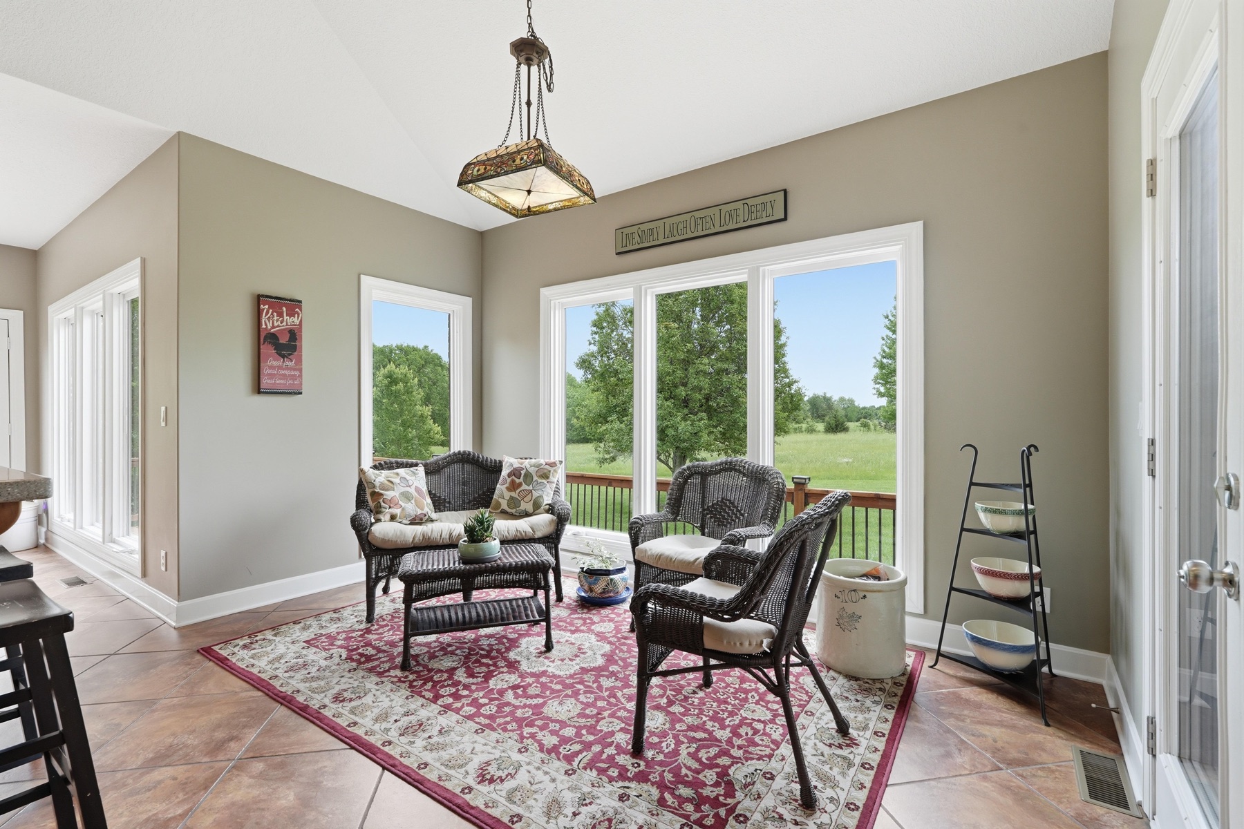 Sun room from a second angle showing the stained-glass pendant lantern hanging at center, wicker chairs on a red Persian rug, two banks of windows on adjacent walls looking out to the green pasture, the sign reading Live Simply Laugh Often Love Deeply above the windows