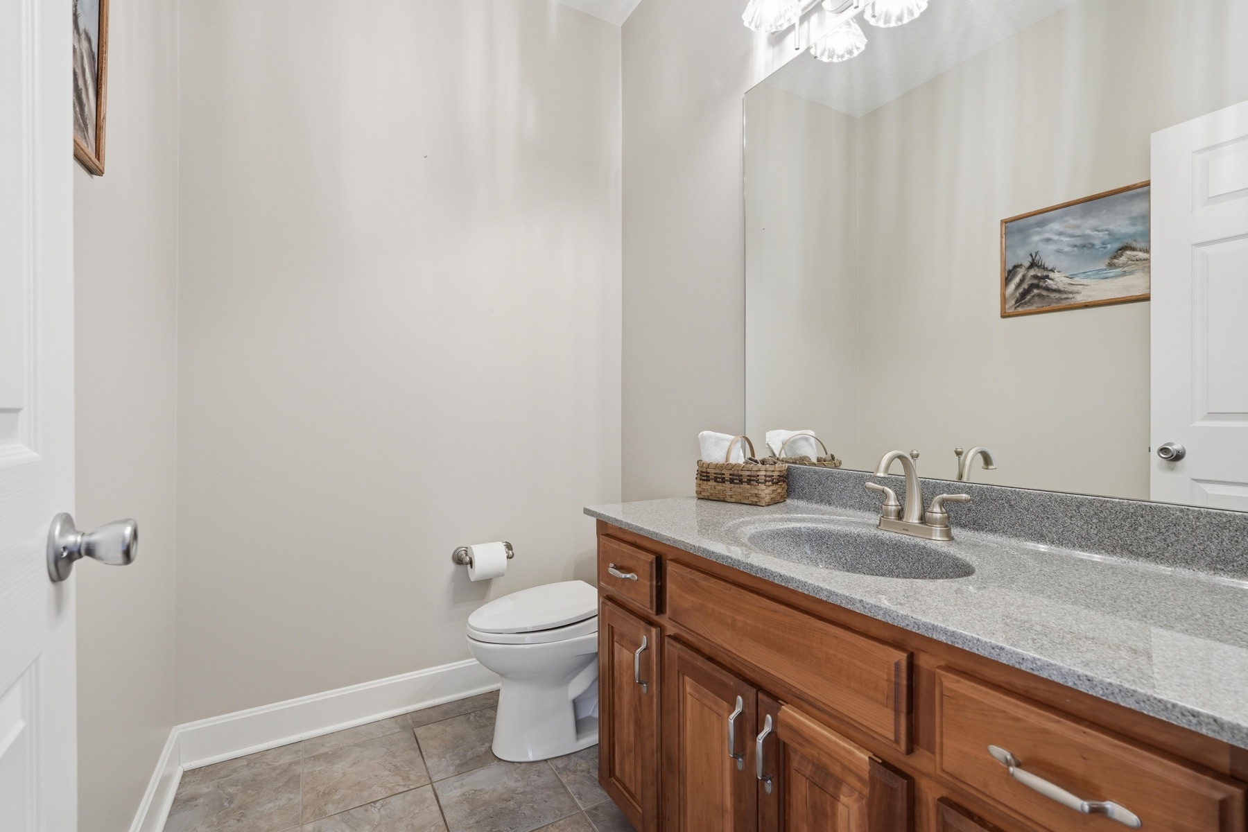 Half bath with a dark cherry-wood vanity, granite countertop, a four-bulb vanity light over a wide mirror, framed coastal artwork above the toilet, and tile floor