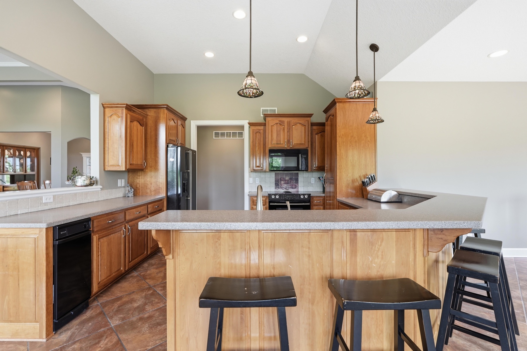 Kitchen wide angle showing the full cherry-wood cabinet wall with stainless French-door refrigerator at center, a black dishwasher, granite countertops, the vaulted ceiling overhead, and three pendant lights hanging over the central island with bar stools