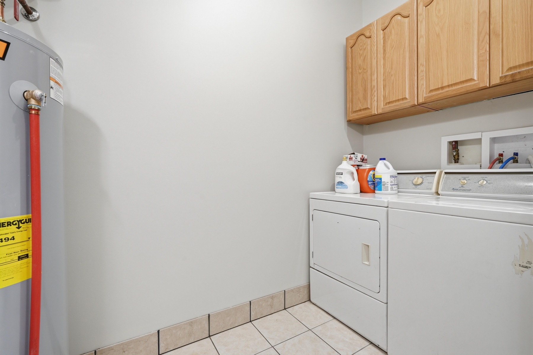 Utility laundry room with an EnergyGuide-tagged water heater on the left, side-by-side white washer and dryer at center, a wall-mounted upper cabinet over the machines for detergent storage, and tile floor underfoot