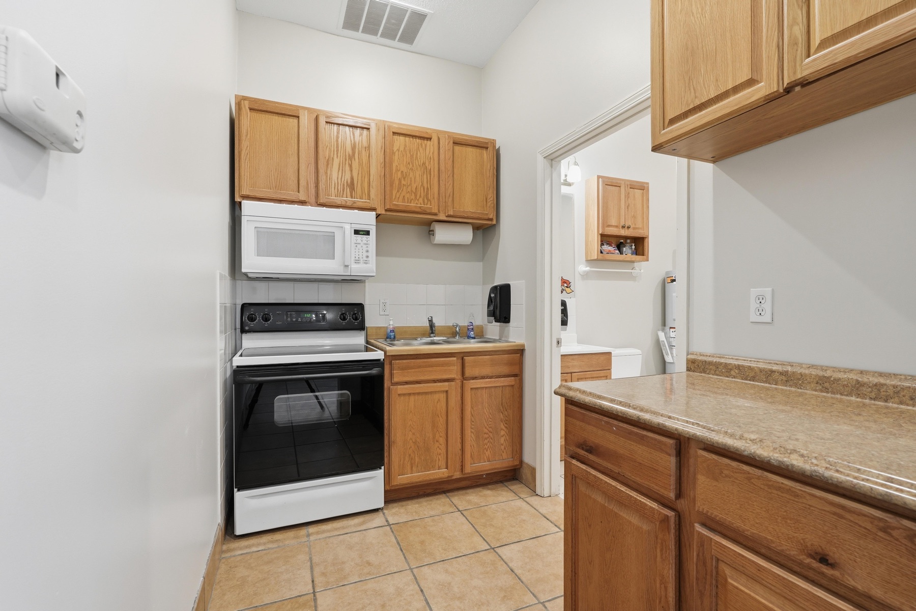 Mezzanine kitchenette inside the shop, oak cabinetry, white electric range with over-the-range microwave, single basin sink, tile backsplash, and an opening to the right leading to a small bathroom and the mezzanine office
