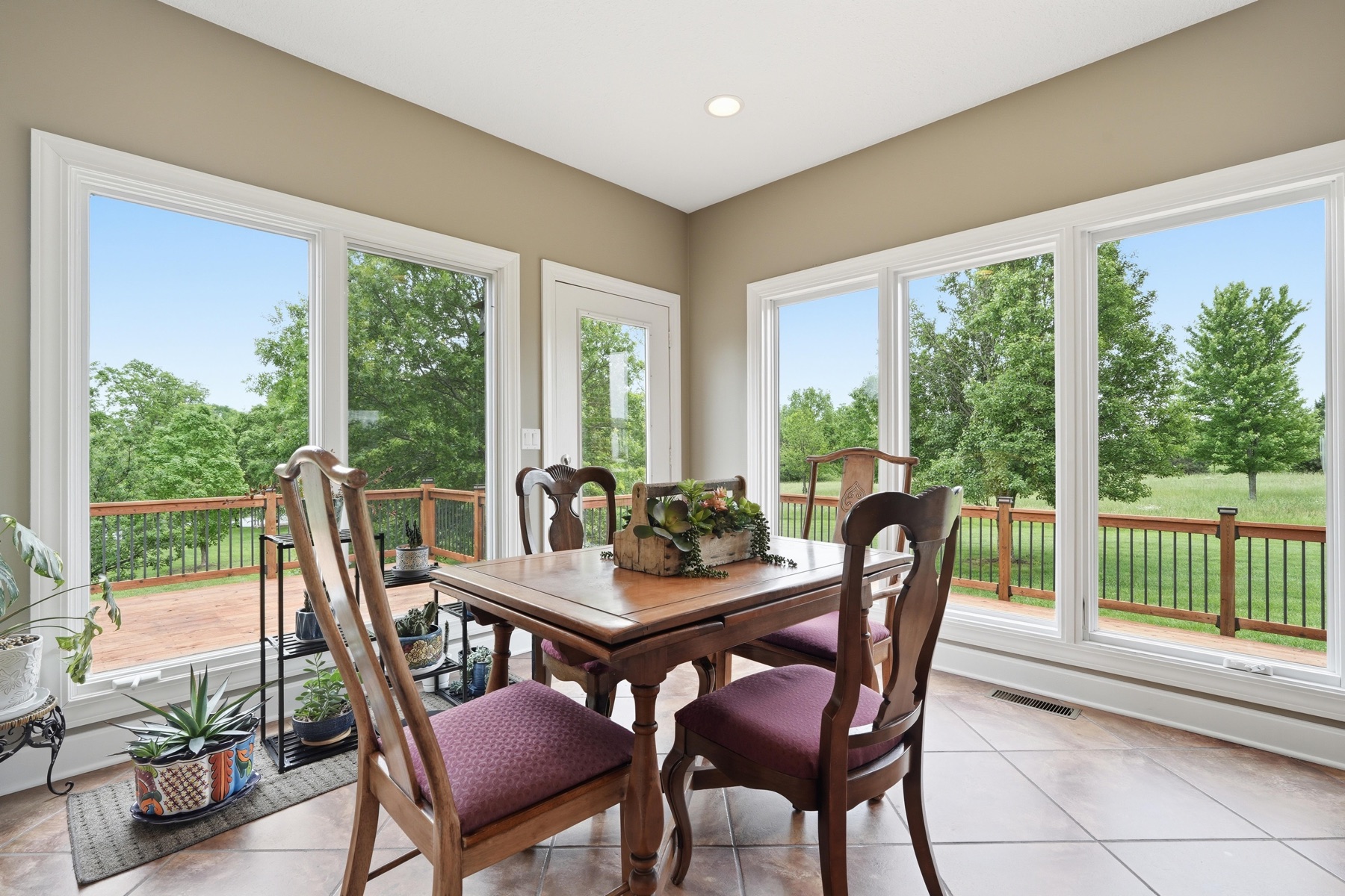 Sun room set up for dining with a small wood table and four high-backed chairs centered on a Persian rug, two banks of tall windows on adjacent walls looking onto the green back lawn, and a metal railing on a deck visible through the glass