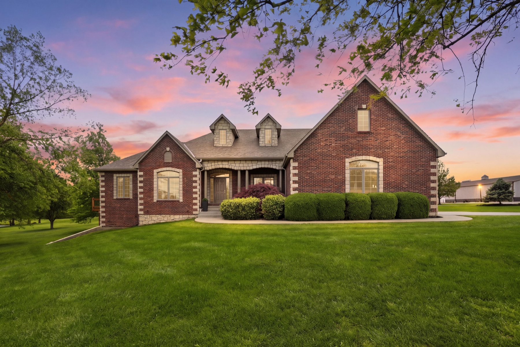 Front elevation of 26619 K-68 Hwy at twilight, all-brick exterior with cedar-shake gable dormers, stone-trimmed entry, manicured hedges along the foundation, a pink and lavender sunset sky behind the home, and a faint silhouette of the commercial shop visible to the right rear