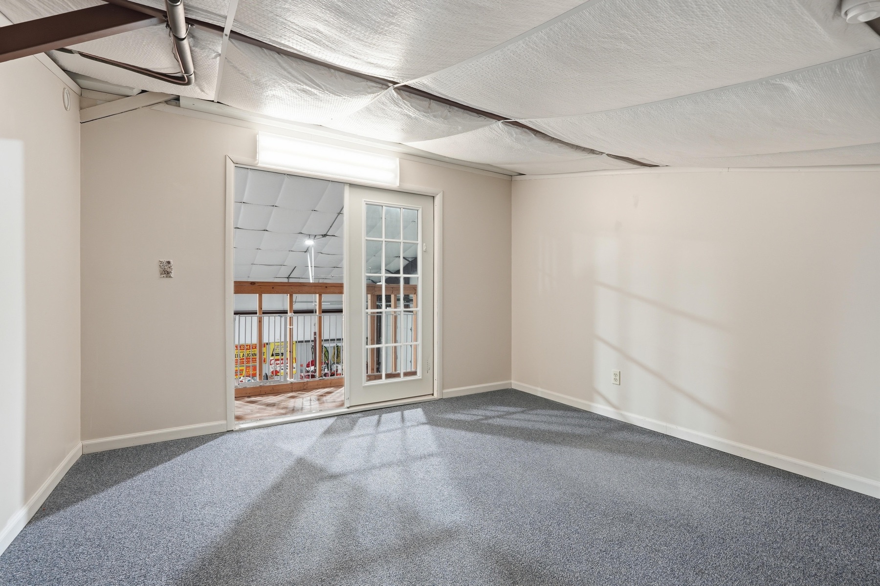 Unfinished basement room with French doors looking through to the finished lower-level area, carpeted floor, exposed insulation overhead, a single fluorescent light fixture, and drywalled walls already finished