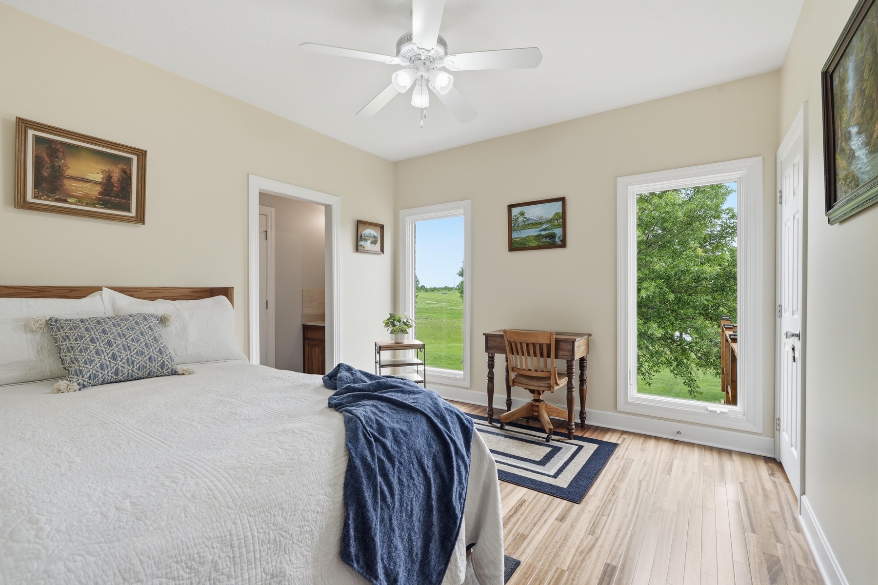 Bedroom with two windows looking out onto green Kansas pasture, a queen bed with white quilt, a small wooden writing desk and chair in the corner, framed prints on the walls, hardwood floors, and a ceiling fan overhead