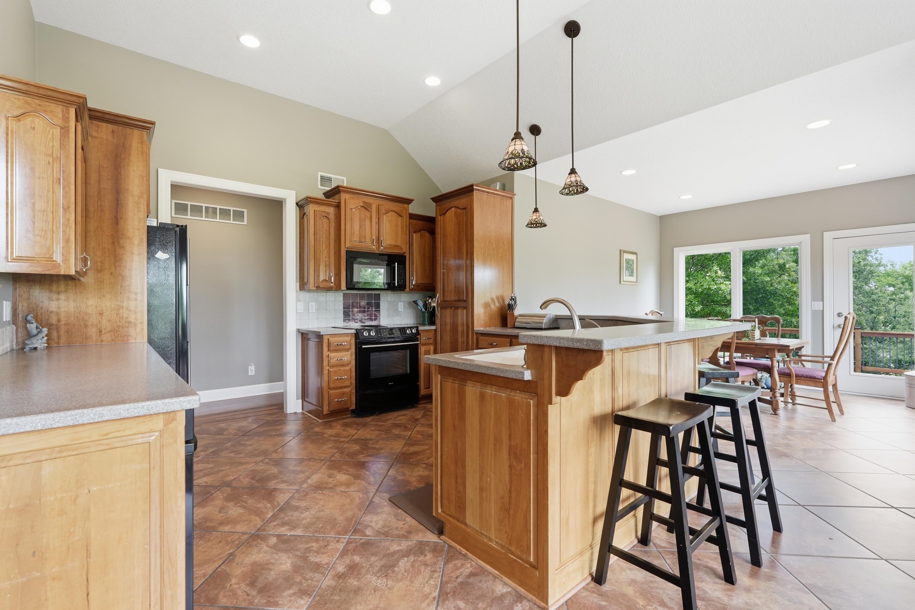 Kitchen looking south showing the long cherry-wood cabinet wall on the left with a black electric range and microwave, an island with three bar stools at center, three copper-and-glass pendant lights hanging overhead, the dining table beyond the island, and large windows letting in pasture light