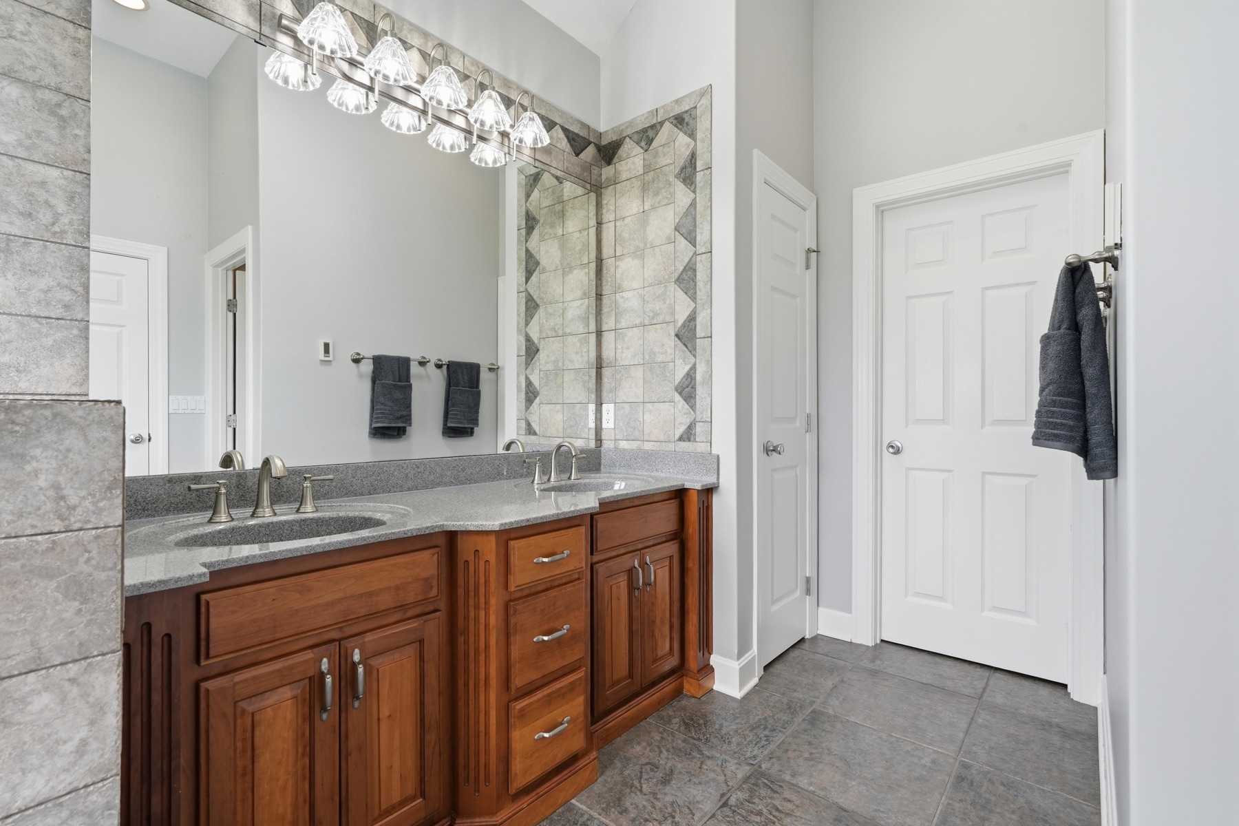 Primary bath cherry double-sink vanity with a long granite countertop, a wide framed mirror reflecting the chevron-tiled shower behind, brushed nickel fixtures, a large bulb vanity light overhead, and a doorway to the closet on the right