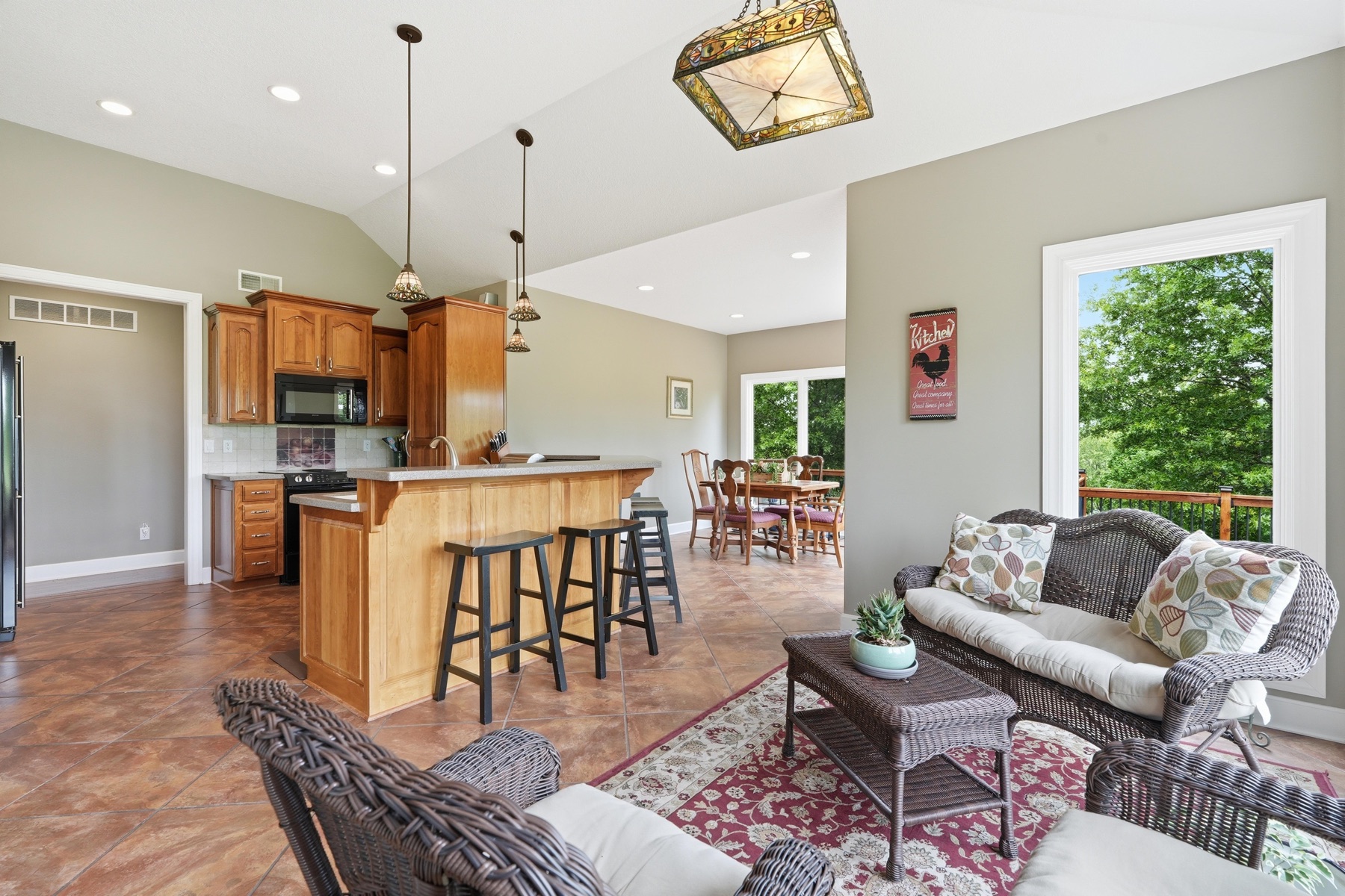Kitchen with a stained-glass pendant fixture over the breakfast peninsula, wicker sitting chairs in the foreground around a coffee table on a red rug, the dining table visible through to the back, and a picture window framing a Rooster sign and the green tree line outside