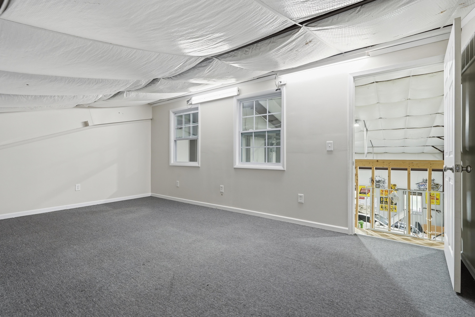 Basement room with carpeting underfoot, two small windows with white grids on the side wall, exposed white insulation on the ceiling, an opening on the right revealing the framing of the next room under construction, and drywalled finished walls otherwise